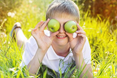 Ein Kind hält sich zwei Äpfel vor die Augen, während es auf einer Wiese liegt.
